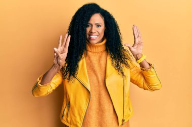 Orta yaşlı Afro-Amerikalı kadın yün kışlık kazak ve deri ceket giyip sekiz numaralı parmaklarıyla işaret ederken kendinden emin ve mutlu gülümsüyor.. 