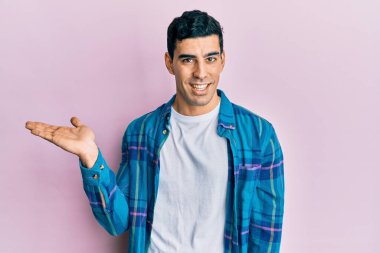 Yakışıklı İspanyol bir adam günlük kıyafetler giymiş neşeli bir şekilde gülümsüyor ve avuç içiyle kameraya bakıyor.. 