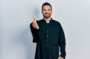 Sakallı yakışıklı, Katolik rahip cübbesi giyen yakışıklı bir adam dostça el sıkışma ve hoş geldin hediyesi olarak gülümsüyor. başarılı iş. 