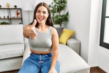 Genç esmer kadın evdeki kanepede oturup dostça gülümseyerek selamlaşma ve selamlama sunuyor. başarılı iş. 