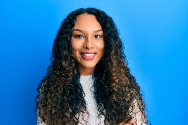 Gündelik kıyafetler giyen, kollarını kavuşturup kameraya gülümseyen genç Latin kadın. pozitif kişi. 