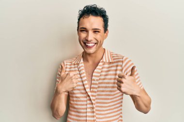 Makyaj yapan yakışıklı bir adam günlük tişört giyer, başarı işareti yapar, el hareketleriyle olumlu bir jest yapar, gülümser ve mutlu olur. Neşeli ifade ve kazanan jest. 
