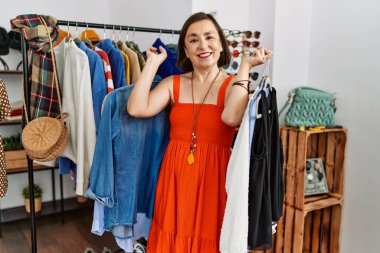 Orta yaşlı İspanyol kadın, mağazada elinde elbise askısıyla gülümsüyor.