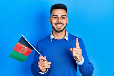 Sakallı, Afgan bayrağı tutan genç İspanyol bir adam bir fikir veya soruyla gülümsüyor ve mutlu bir yüzle işaret ediyor. 