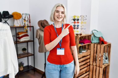 Young caucasian woman working as manager at retail boutique cheerful with a smile of face pointing with hand and finger up to the side with happy and natural expression on face 