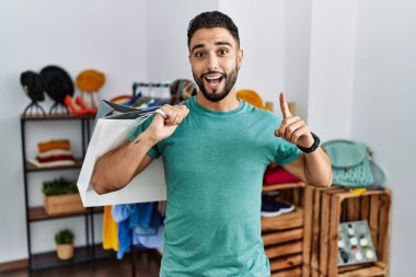 Satış mağazasında sakallı, alışveriş torbaları tutan genç yakışıklı adam şaşırmış, şaşırmış ve parmaklarıyla işaret ediyor ve kollarını kaldırıyordu.. 
