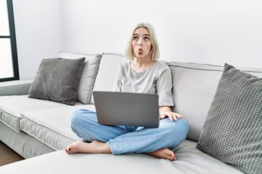 Young caucasian woman using laptop at home sitting on the sofa making fish face with lips, crazy and comical gesture. funny expression. 