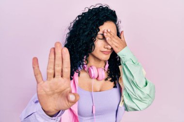 Young hispanic woman with curly hair wearing gym clothes and using headphones covering eyes with hands and doing stop gesture with sad and fear expression. embarrassed and negative concept. 
