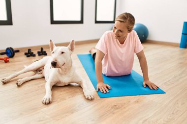 Genç beyaz kadın gülümsüyor spor merkezinde köpekle esniyor.