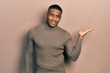 Balıkçı yaka kazak giyen, neşeli gülümseyen, avuç içi kameraya bakan genç siyahi bir adam.. 