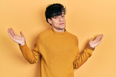 Yakışıklı hippi genç adam gündelik sarı süveter giyiyor ve elleri ve kolları havada kafası karışık bir ifade takınıyor. şüphe kavramı. 