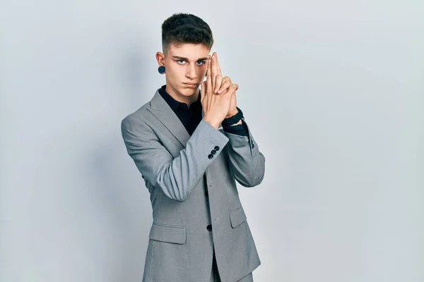 Young caucasian boy with ears dilation wearing business jacket holding symbolic gun with hand gesture, playing killing shooting weapons, angry face