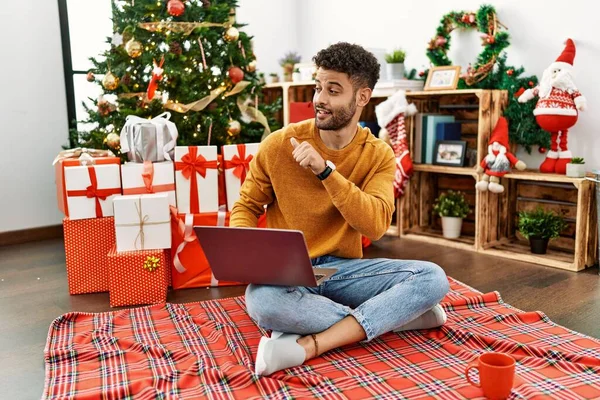 청년은 크리스마스 앉아서 노트북을 사용하며 자신있게 웃으면서 손가락을 가리키고 있습니다 — 스톡 사진