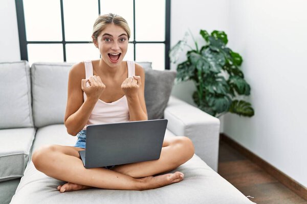 Молодая блондинка, сидя дома на диване, радовалась и удивлялась успеху с поднятыми руками и открытыми глазами. победитель концепция. 