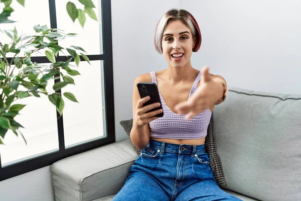 ソファに座って挨拶や歓迎として握手を提供優しい笑みを浮かべてスマートフォンの入力メッセージを使用して若い美しい女性 成功したビジネス — ストック写真