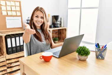 Young brunette woman working at the office with laptop smiling friendly offering handshake as greeting and welcoming. successful business. 