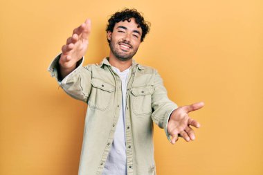 Gündelik giysiler giymiş genç İspanyol adam kameraya bakıp sarılmak için kollarını açmış gülümsüyor. Mutluluğu kucaklayan neşeli bir ifade.. 