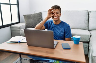 Young handsome hispanic man using laptop sitting on the floor doing ok gesture with hand smiling, eye looking through fingers with happy face. 