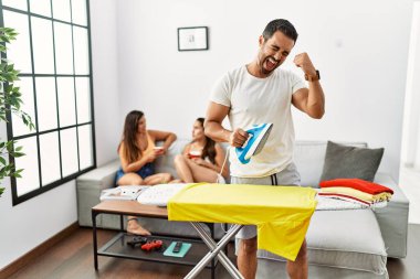 Genç İspanyol adam evde kıyafetlerini ütülüyor. Çok mutlu ve heyecanlı. Kazanan jest yapıyor, kolları havada, gülümsüyor ve başarı için bağırıyor. kutlama konsepti. 