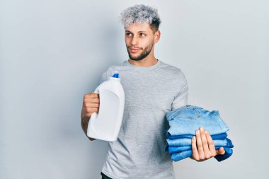Modern boyalı saçlı, çamaşırhane için kot pantolon ve deterjan şişesi tutan genç İspanyol bir adam. Gülümsüyor ve başka tarafa bakıyor.. 