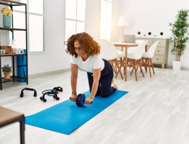 Orta yaşlı İspanyol kadın gülümsüyor. Karın kaslarını çalıştırırken evde tekerlekli sandalye kullanıyor.