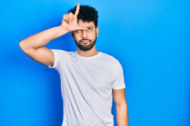 Young arab man with beard wearing casual white t shirt making fun of people with fingers on forehead doing loser gesture mocking and insulting. 