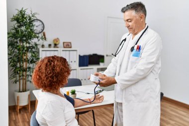 Orta yaşlı erkek ve kadın doktor üniforması giyiyorlar. Tansiyon ölçer kullanıyorlar. Klinikte tıbbi konsültasyon yapıyorlar.