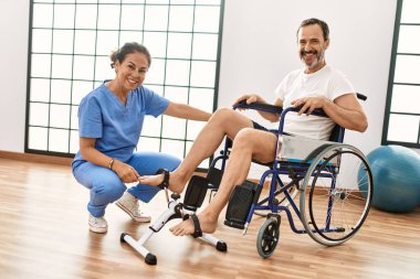 Orta yaşlı bir adam ve kadın fizik tedavi kliniğinde tekerlekli sandalyede pedal çeviriyorlar.