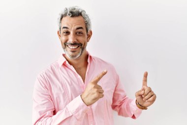 Middle age hispanic man standing over isolated background smiling and looking at the camera pointing with two hands and fingers to the side. 