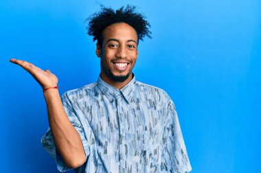 Sakallı, sıradan giyinmiş, neşeli gülümseyen, avuç içi dolu bir şekilde kameraya bakan Afro-Amerikalı genç bir adam.. 