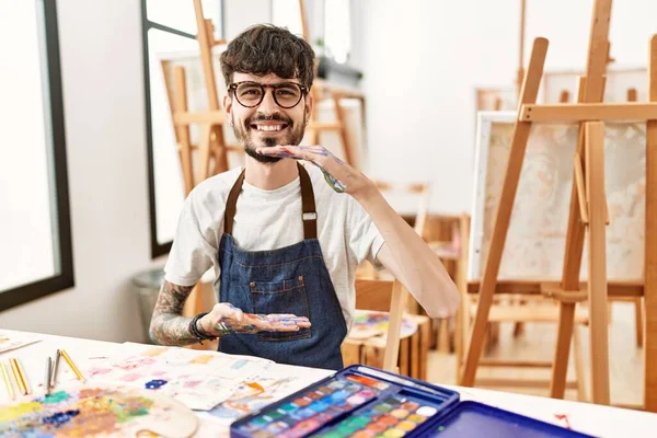 Hispanic man with beard at art studio gesturing with hands showing big and large size sign, measure symbol. smiling looking at the camera. measuring concept.