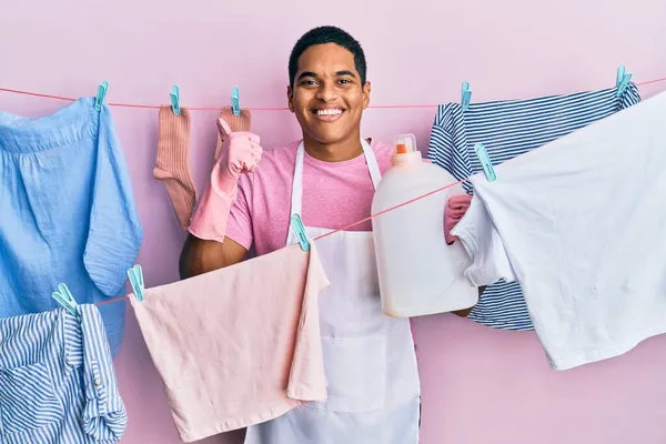 Jovem Homem Hispânico Bonito Fazendo Lavanderia Segurando Garrafa Detergente Sorrindo — Fotografia de Stock