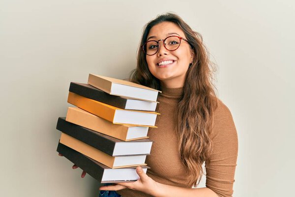 Young hispanic girl holding a pile of books smiling with a happy and cool smile on face. showing teeth. 