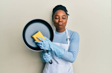 Saçları örülmüş, önlük takmış, elinde çamaşır makinesi olan Afro-Amerikalı bir kadın kameraya bakıp öpüşüyor ve çok seksi oluyor. aşk ifadesi. 