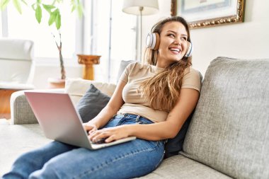 Laptop ve kulaklık kullanan genç İspanyol kız evdeki koltukta oturuyor..