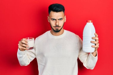 Sakallı, elinde bir bardak süt ve plastik şişe tutan, kuşkucu ve gergin bir İspanyol genç, sorun yüzünden surat asıyor. negatif kişi. 