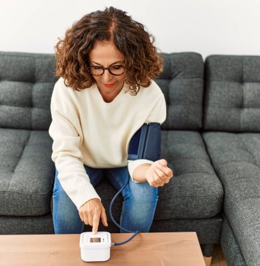 Kan basıncı monitörü kullanan olgun İspanyol kadın evdeki kanepede oturuyor.