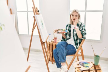 Young artist woman painting on canvas at art studio looking stressed and nervous with hands on mouth biting nails. anxiety problem. 