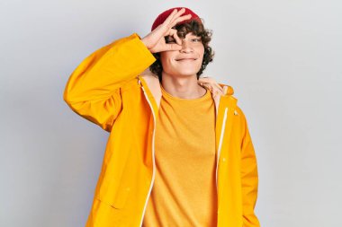 Handsome young man wearing yellow raincoat smiling happy doing ok sign with hand on eye looking through fingers 