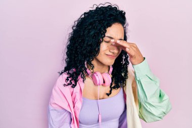 Young hispanic woman with curly hair wearing gym clothes and using headphones tired rubbing nose and eyes feeling fatigue and headache. stress and frustration concept. 