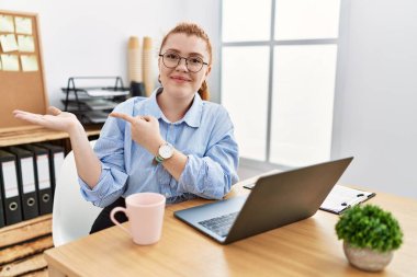 Ofiste çalışan genç kızıl saçlı kadın bilgisayarını kullanarak kameraya hayret ediyor ve gülümsüyor.. 