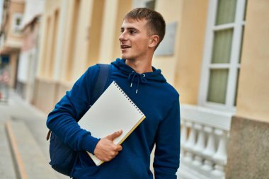 Üniversitede elinde defterle gülümseyen sarışın genç bir öğrenci..