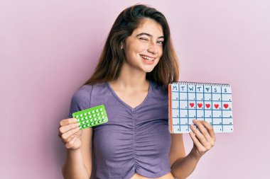 Elinde doğum kontrol hapları ve regl takvimi olan beyaz bir kadın kameraya seksi bir ifadeyle bakıyor, neşeli ve mutlu bir yüzle.. 