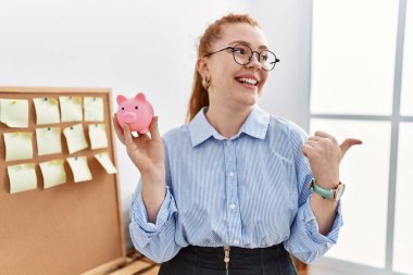 Ofiste domuz kumbarasını tutan genç kızıl saçlı kadın başparmağıyla yan tarafı işaret ediyor ve ağzı açık gülümsüyor. 