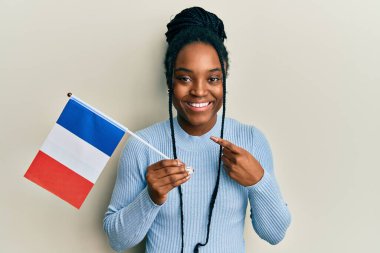 Saçları örgülü Afrika kökenli Amerikalı bir kadın Fransa bayrağı tutarken mutlu bir şekilde el ve parmak işareti yapıyor. 