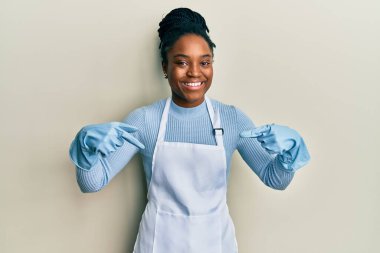 Saçları örgülü, daha temiz önlüklü ve eldivenli, kendine güvenen, gülümseyen, kendini gururlu ve mutlu parmaklarıyla gösteren Afrikalı Amerikalı bir kadın.. 