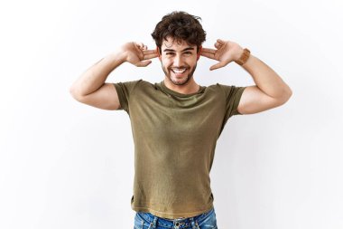 Hispanic man standing over isolated white background smiling pulling ears with fingers, funny gesture. audition problem 