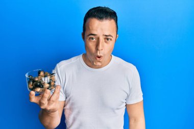 Handsome young man holding raw hazelnuts scared and amazed with open mouth for surprise, disbelief face 