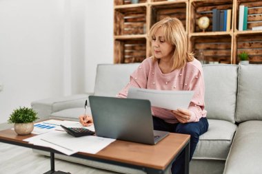 Orta yaşlı sarışın kadın evdeki kanepede oturarak tele-çalışmaya odaklanmış..