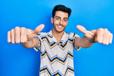 Latin asıllı genç adam gündelik giysiler giyip olumlu jestler yapıyor. Elleriyle gülümsüyor ve başarı için seviniyor. kazanan jest. 
